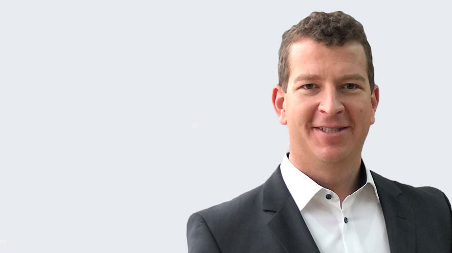 Ralph-Jürgen Detering: Porträt eines leicht lächelnden Mannes im dunklen Anzug mit weißem Hemd vor hellgrauem Hintergrund.