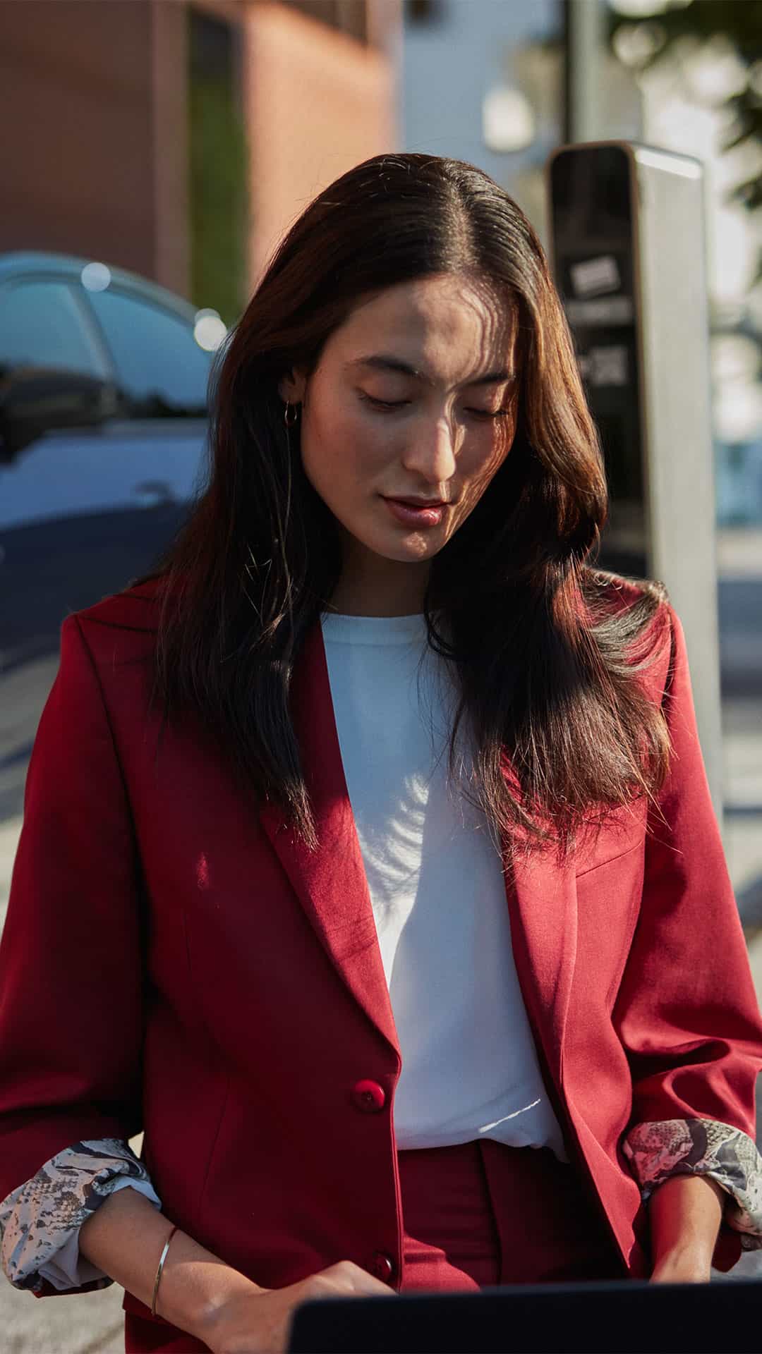 Frau in rotem Anzug arbeitet draußen am Laptop, im Hintergrund steht ein blauer Audi auf einer städtischen Straße.
