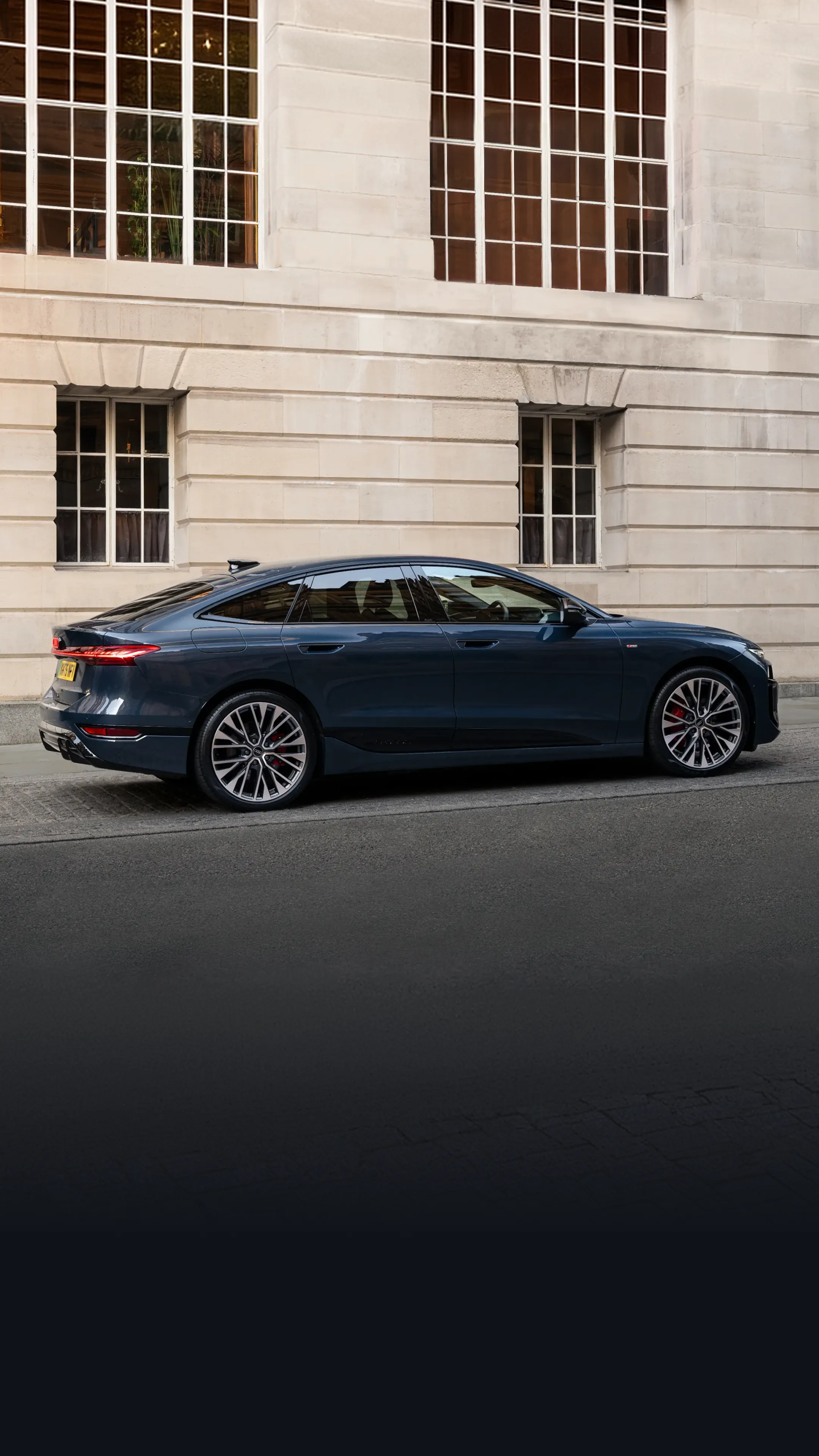 Dark blue Audi Sportback e-tron parked on a city street, shown in full side profile against a stone building facade. The vehicle features a sleek fastback roofline, large multi-spoke alloy wheels, red brake callipers and slim rear LED lights. Natural daylight highlights the aerodynamic body contours and premium design details.