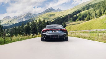 Dunkelgrauer Audi RS e-tron GT performance (Heckansicht) fährt auf einer Bergstraße, im Hintergrund grüne Hügel und schneebedeckte Gipfel.