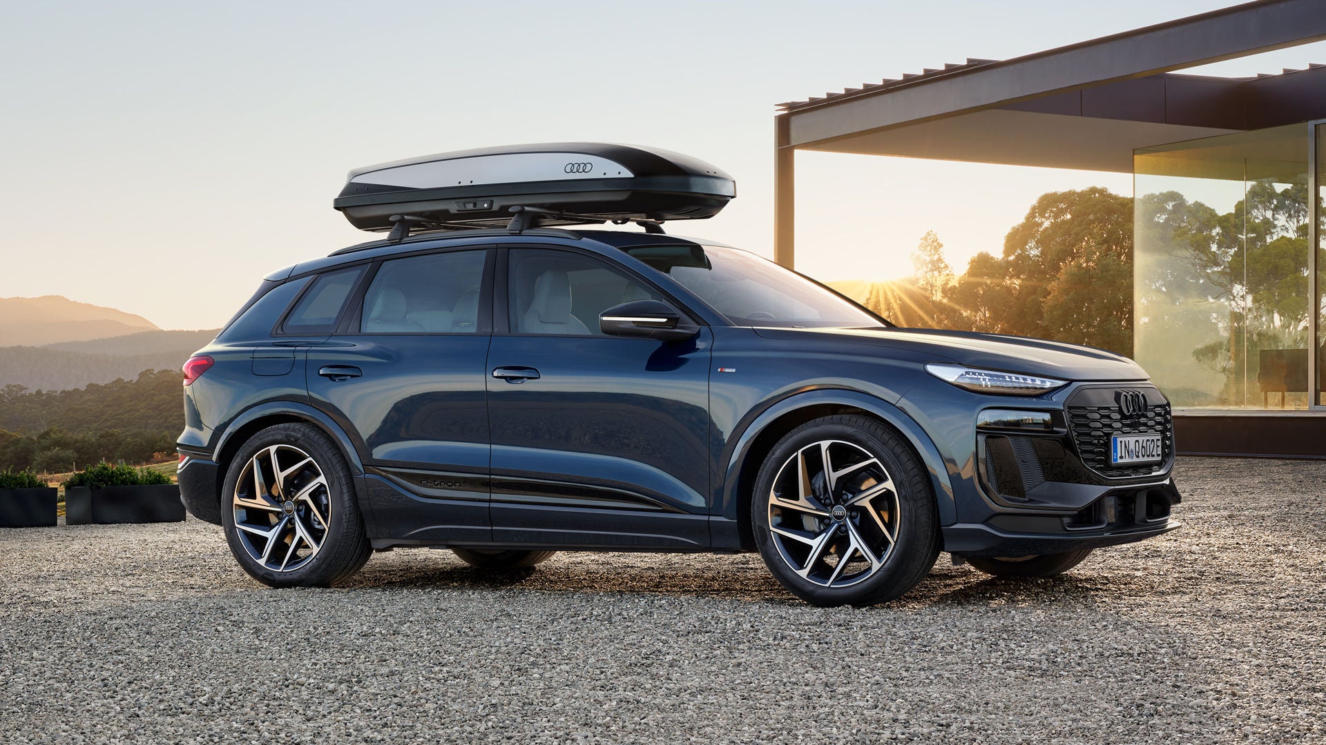 De Q6 e-tron met dakkoffer staat dwars geparkeerd op het grindterrein voor een modern huis, verlicht door de ondergaande zon.