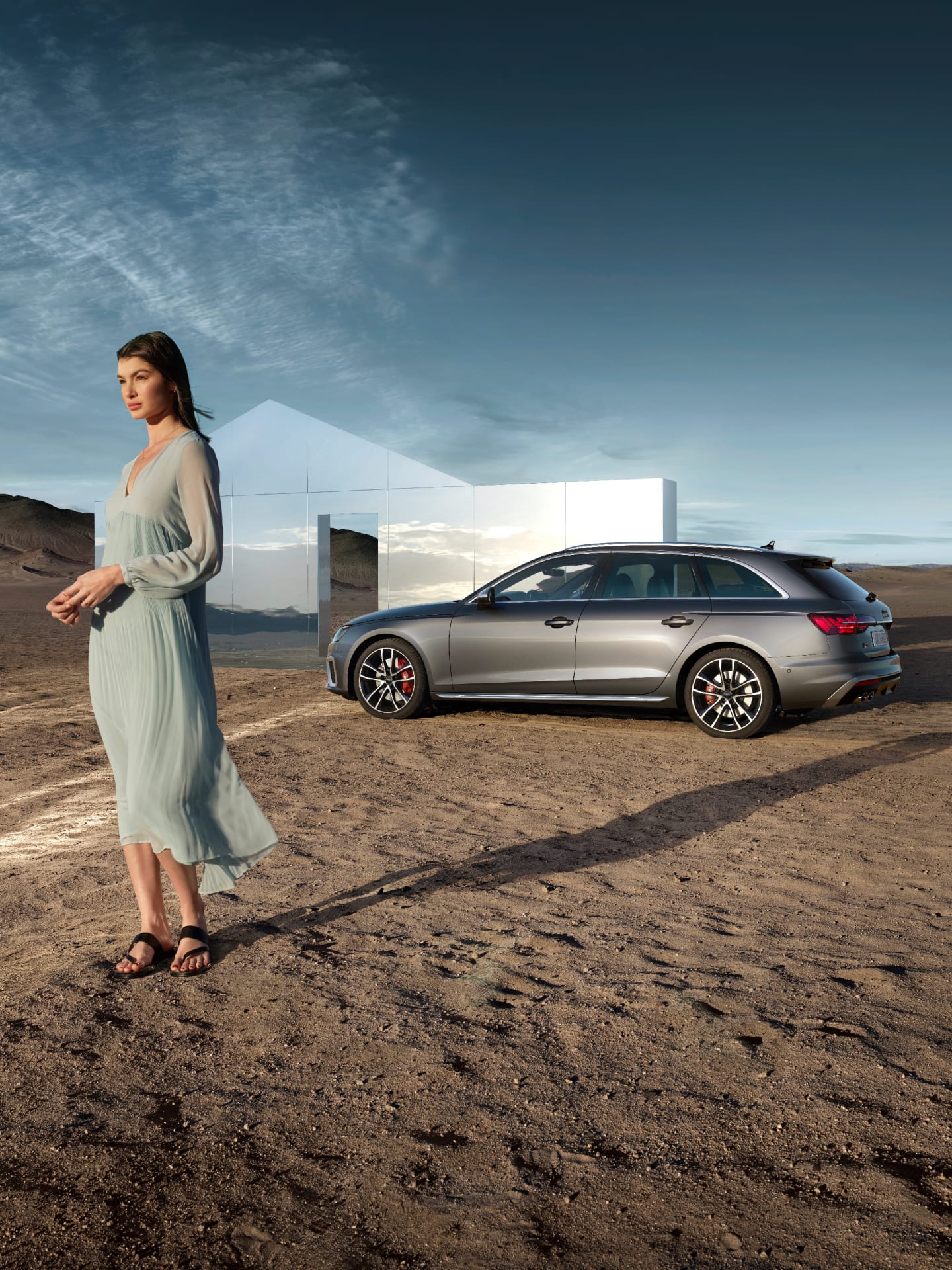 A person in a long dress stands by a modern car outside a glass house in a desert.