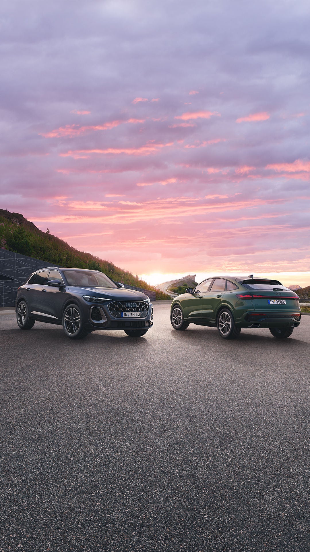 Deux Audi Q5 sont garées sur une route de montagne au coucher du soleil. Le ciel est peint de couleurs douces et des collines verdoyantes sont visibles en arrière-plan.