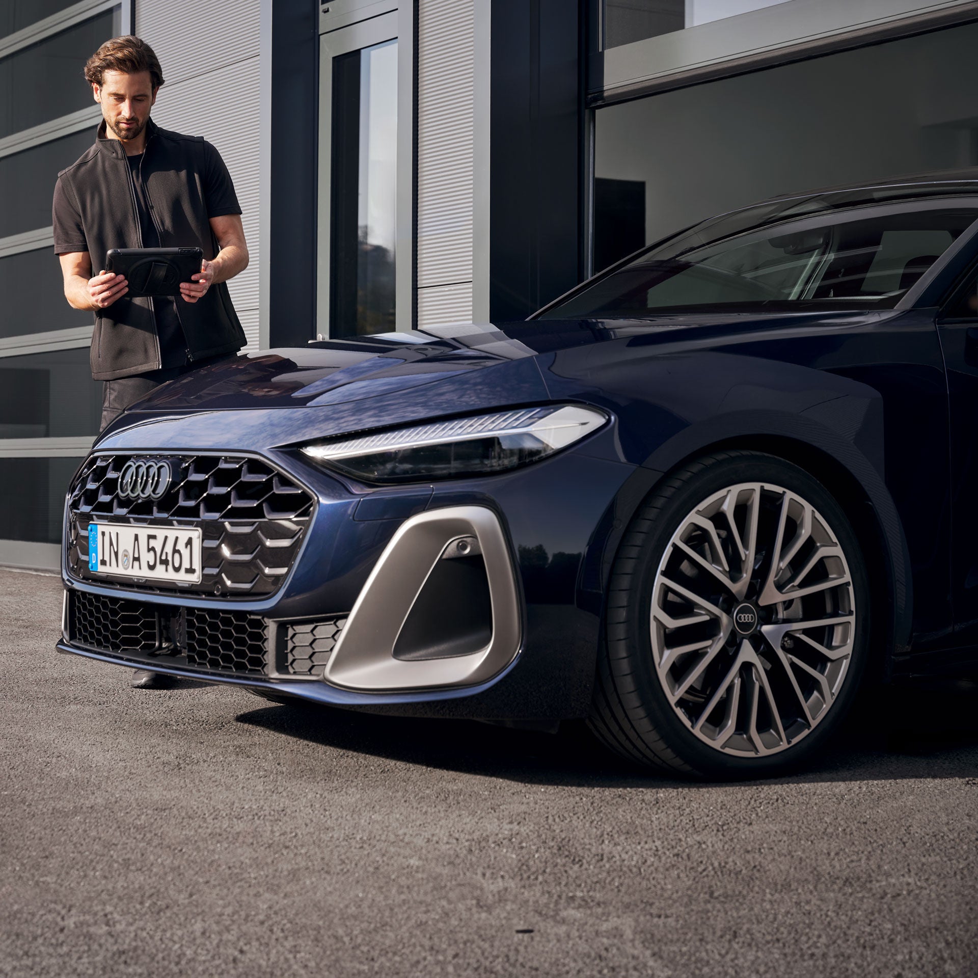 Mann steht neben einem dunkelblauen Audi und bedient ein Tablet vor einer Werkstatt.