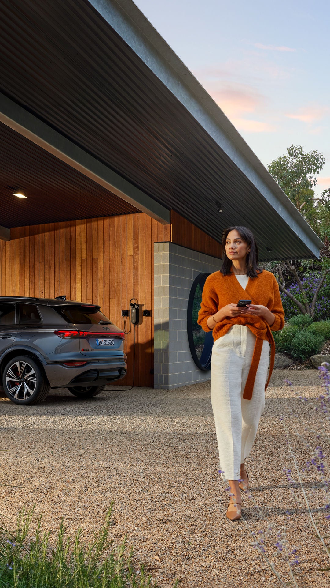 Dunkelhaarige Frau mit Smartphone läuft vor einem geparkten, ladenden Audi Q6 SUV e-tron unter Carport mit Holzfassade bei Tageslicht.