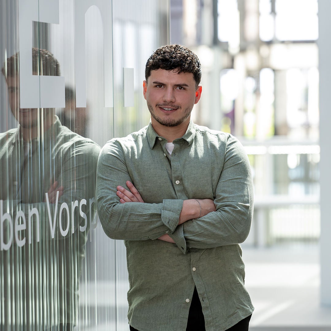 Ergon ist mit verschränkten Armen an eine Wand gelehnt und lächelt in die Kamera