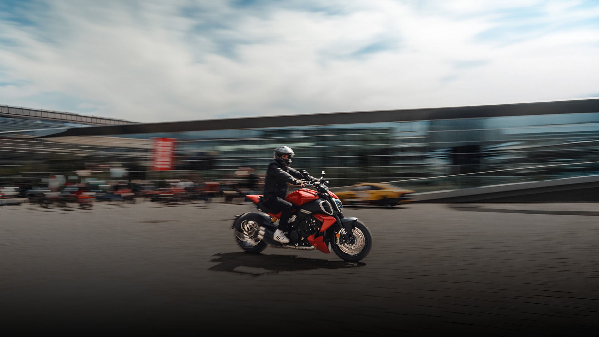 Motorradfahrer fährt dynamisch über Platz vor modernem Gebäude, Hintergrund mit Bewegungsunschärfe.