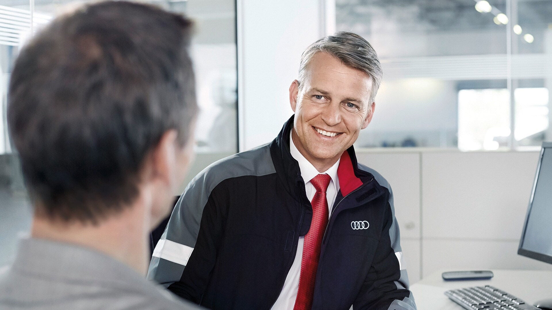 Mitarbeiter im Audi-Outfit mit roter Krawatte lächelt einem Kunden an einem Schreibtisch freundlich zu.