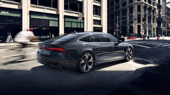 Ein grauer Audi A7 fährt durch eine Straßenecke in der Stadt, Fahrradfahrer unscharf im Vordergrund, Gebäude im Hintergrund.