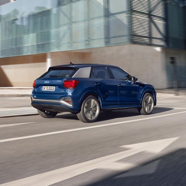 Ein blauer Audi Q2 fährt dynamisch durch die Stadt, modernes Gebäude mit Glasfassade im Hintergrund.