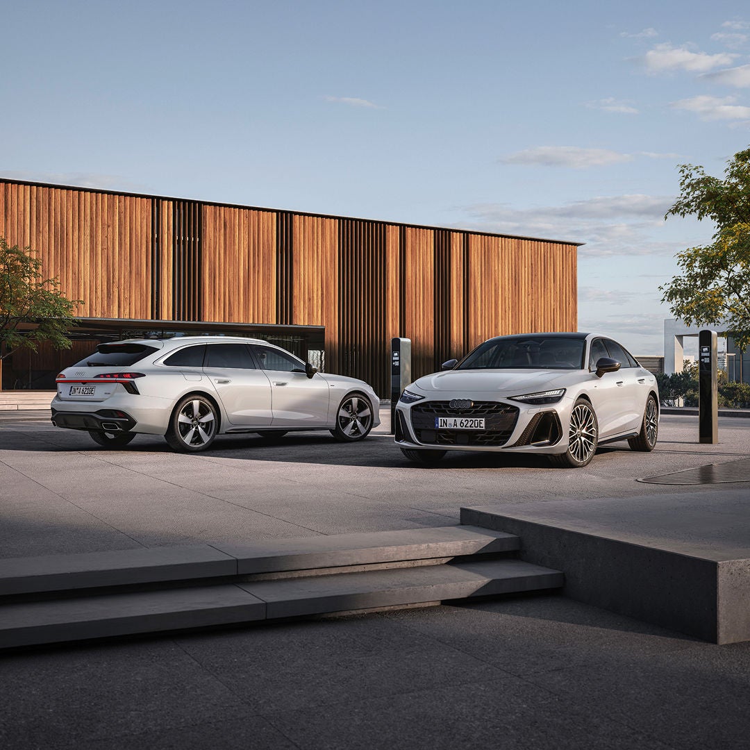 A white Audi and a black Audi parked outside a modern wooden building under a clear sky.
