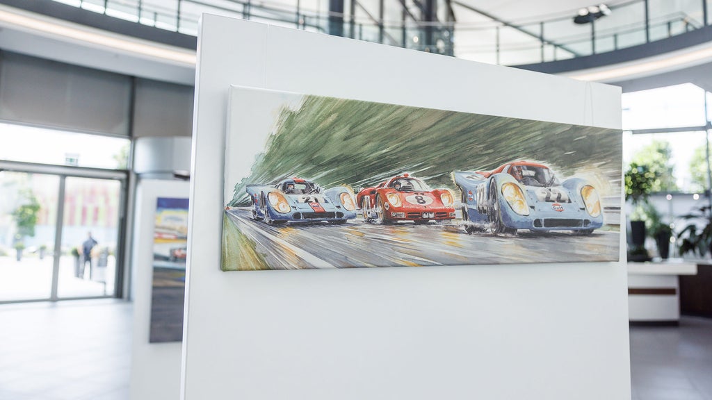 A painting of three race cars in blue and red on a racetrack, displayed on a white backing in a modern lobby.