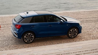 A blue SUV is parked on a cobblestone surface near a waterfront. The scene conveys a calm and serene atmosphere with gentle water in the background.