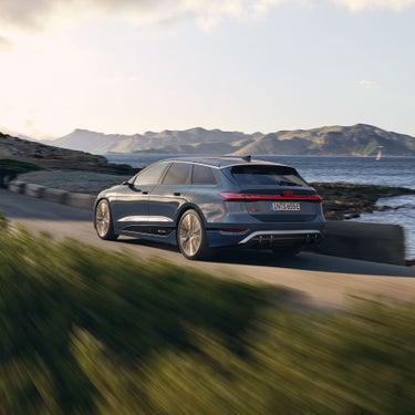 Dunkelblauer Audi S6 Avant e-tron fährt auf Küstenstraße bei Tageslicht, im Hintergrund Berge und Meer.
