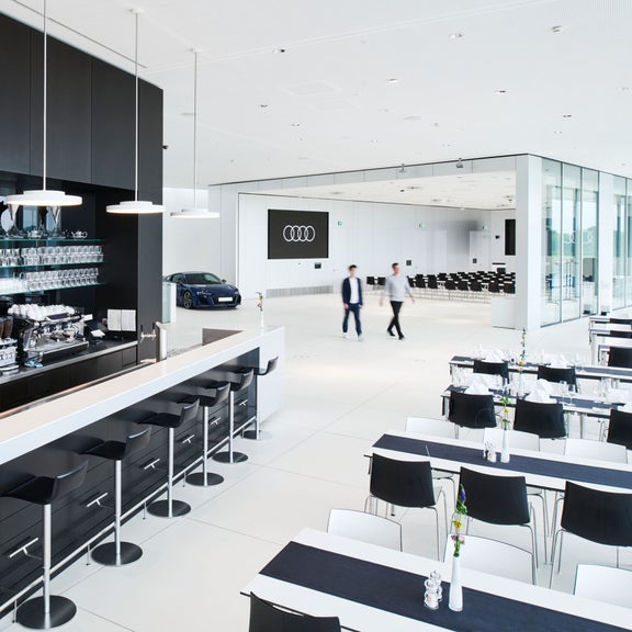 Moderner Veranstaltungsbereich in der Audi drivers lounge mit Bar, Tischen und Blick auf eine Halle mit Audi Präsentationsfläche.