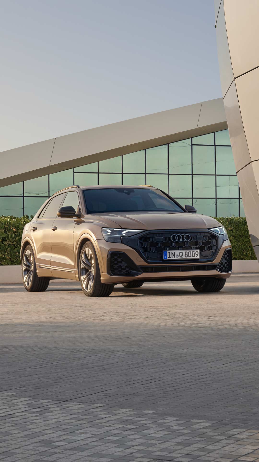 Ein goldener Audi Q8 SUV steht vor einem Gebäude mit Glasfassade und Pflanzen im Hintergrund.