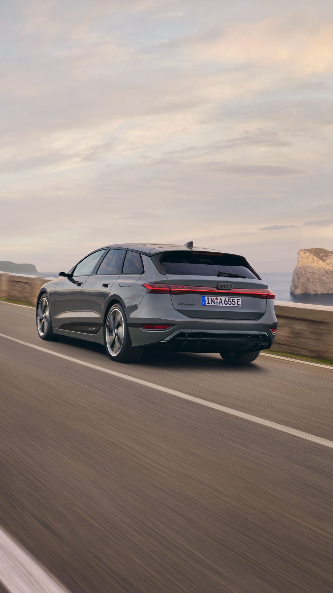 Schräge Heckaufnahme des A6 Avant e-tron, welcher auf einer Straße entlang des Meeres fährt. Im Hintergrund sind Felsen und Berge zu erkennen.