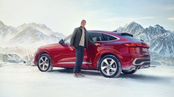 Person steht lächelnd neben einem roten SUV von Audi in verschneiter Berglandschaft.