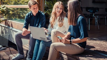 3 junge Menschen sitzen auf Holzstufen und unterhalten sich. Die Frau in der Mitte hat einen Laptop mit Audi Logo auf dem Schoß.