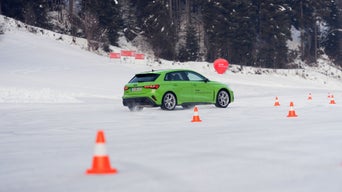 Hellgrüner Audi RS 3 Sportback fährt auf schneebedeckter Strecke zwischen orangen Pylonen, im Hintergrund verschneiter Wald.