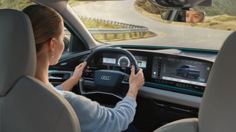 Detailaufnahme des Innenraum-Cockpits eines Audi, von schräg hinter dem Fahrersitz aufgenommen. Eine Frau sitzt am Steuer. Sie blickt auf eine kurvige Straße. Ihre Augen sind im Rückspiegel zu erkennen.