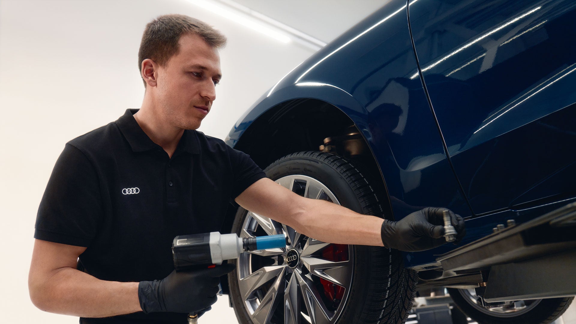 Audi Mitarbeiter in schwarzem Poloshirt montiert ein Rad an einem blauen Auto in der Werkstatt.