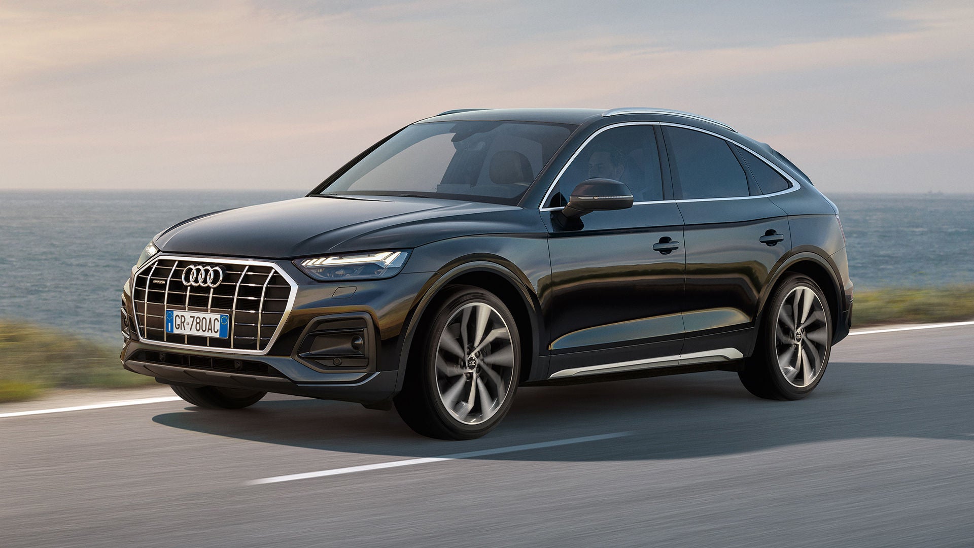  Vista di un uomo alla guida un veicolo Audi che percorre una strada, con mare sullo sfondo.