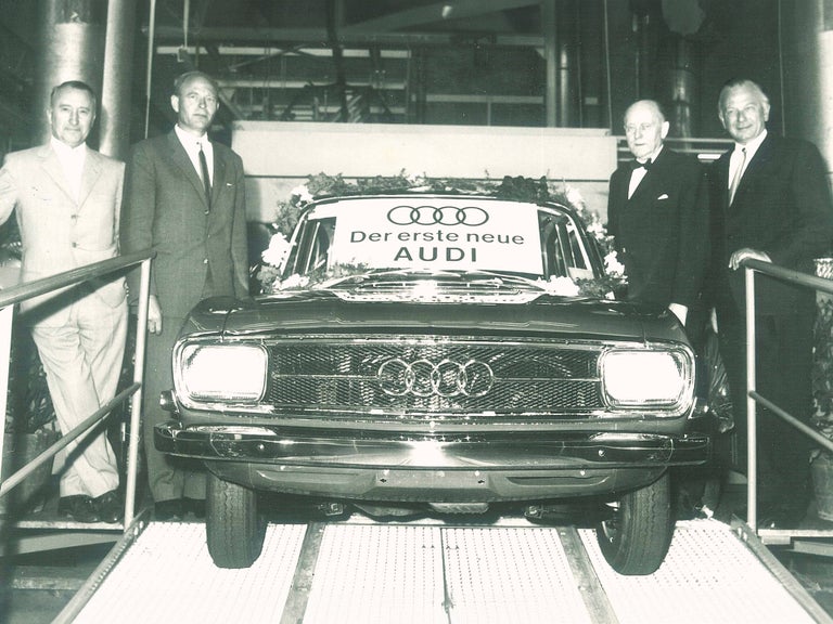 Schwarz-weiß-Aufnahme von mehreren Personen neben einem Auto Union Audi mit einem Schild "Der erste neue AUDI"