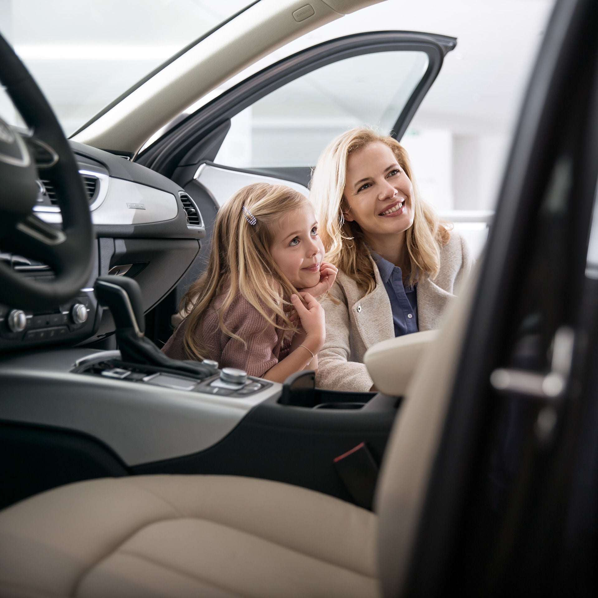 Lächelnde Frau mit kleinem Mädchen, beide sitzen nebeneinander im geöffneten Auto und schauen gemeinsam nach ins Auto.