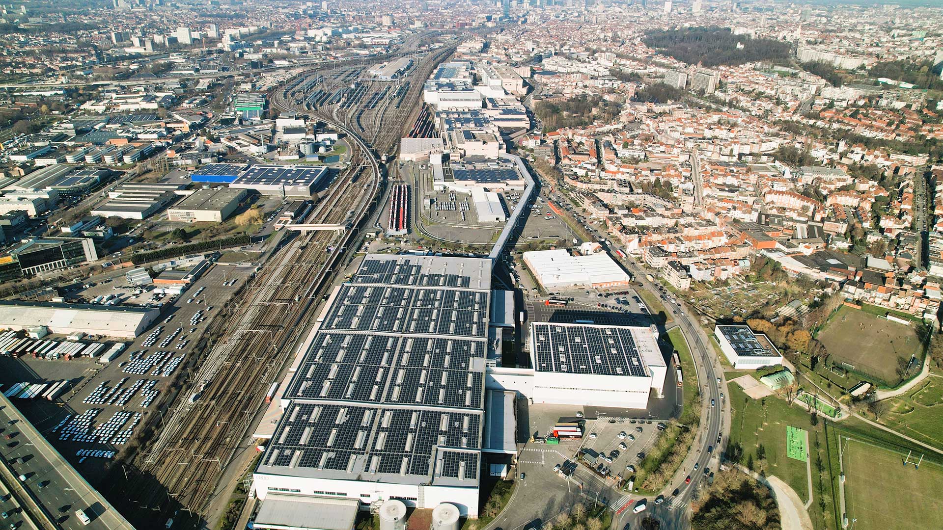Vue aérienne du site de l'usine Audi Brussels. Au premier plan, on aperçoit un grand bâtiment d'usine dont le toit est recouvert de panneaux solaires. Les voies ferrées sont visibles sur la gauche.