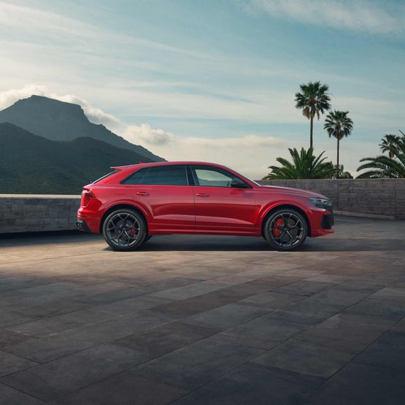 Seitenansicht eines roten Audi RS Q8 SUV performance auf heller Steinfläche vor Berglandschaft und Palmen bei Tageslicht.