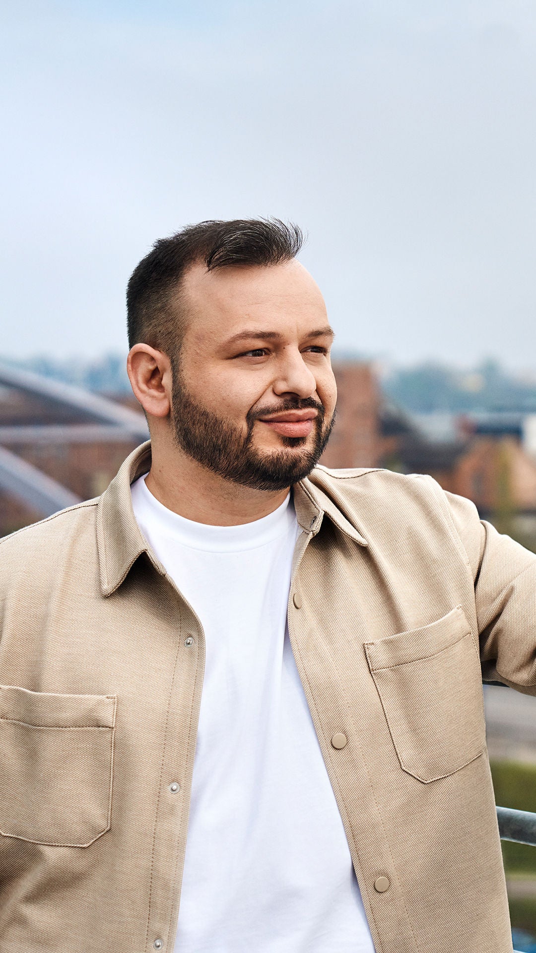 Buenyamin looks to the right, leans against a railing and has a city behind him