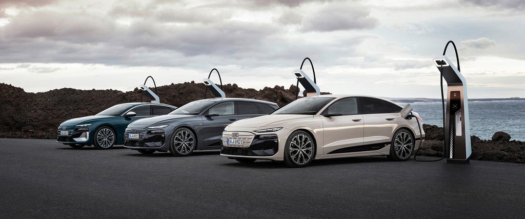 Drei Audi Q6 e-tron Fahrzeuge in verschiedenen Farben stehen an einer Ladestation vor einer felsigen Küstenlandschaft mit bewölktem Himmel. Das vorderste Fahrzeug ist an die Ladesäule angeschlossen.