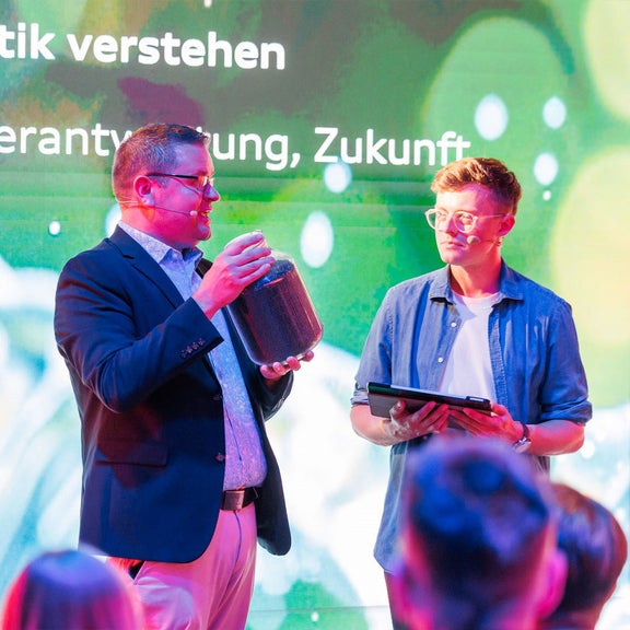 Mann im Sakko hält großes Glas mit Kunststoffgranulat, neben ihm steht ein junger Mann mit Tablet.