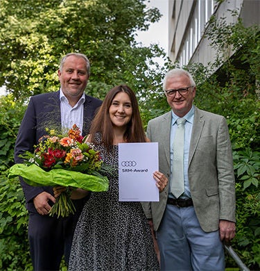 Drei Personen posieren mit einer Auszeichnung und einem Blumenstrauß.