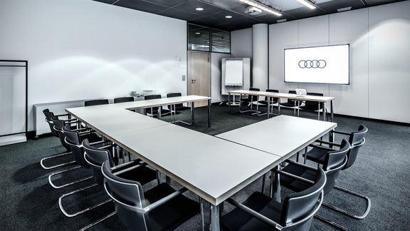 Heller Konferenzraum mit U-förmigem Tisch, schwarzen Stühlen und Audi-Logo auf einem Bildschirm.
