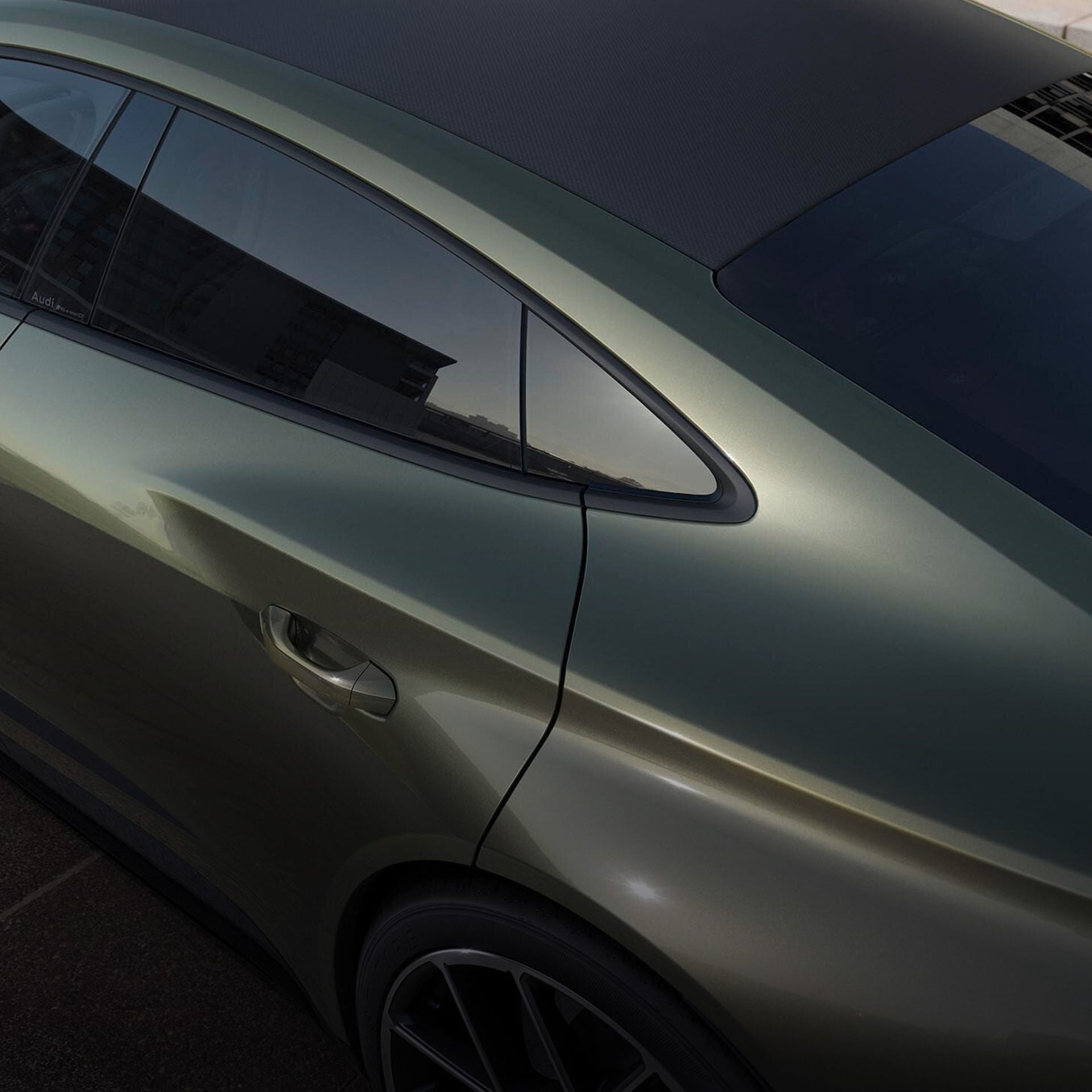 Detailaufnahme eines olivgrünen Audi RS e-tron GT performance mit betont aerodynamischem Design. Zu sehen ist die hintere Tür, das dunkel getönte Seitenfenster und der geschwungene Übergang zur C-Säule. Das Fahrzeug hat bündige Türgriffe und reflektierende Metallic-Lackierung.