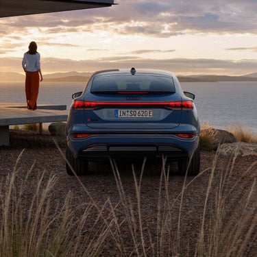 Rückansicht des blauen Audi SQ6 Sportback e-tron vor abendlicher Seelandschaft, Frau steht auf Terrasse und blickt in die Ferne.