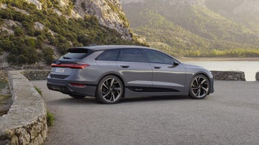 Der A6 Avant e-tron steht seitlich auf einem Kiesparkplatz vor einem See. Im Hintergrund sind von Bäumen bewachsene Berge zu sehen.