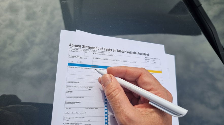 Hand mit Kugelschreiber füllt ein Formular zum Kfz-Unfallbericht auf der Motorhaube eines Autos aus.