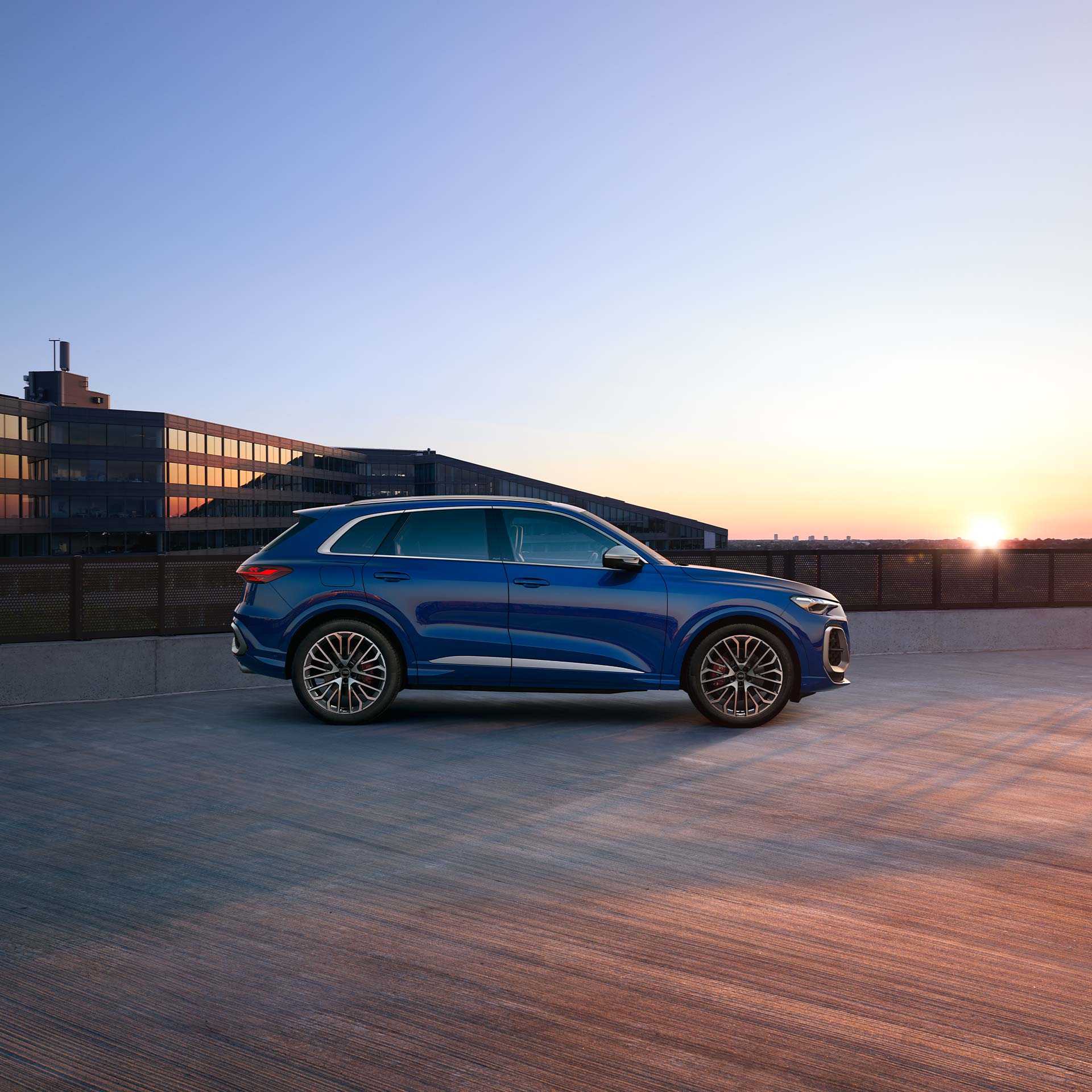 Ein blauer Audi SQ5 SUV seitlich auf einem Parkdeck bei Sonnenuntergang, modernes Bürogebäude im Hintergrund.