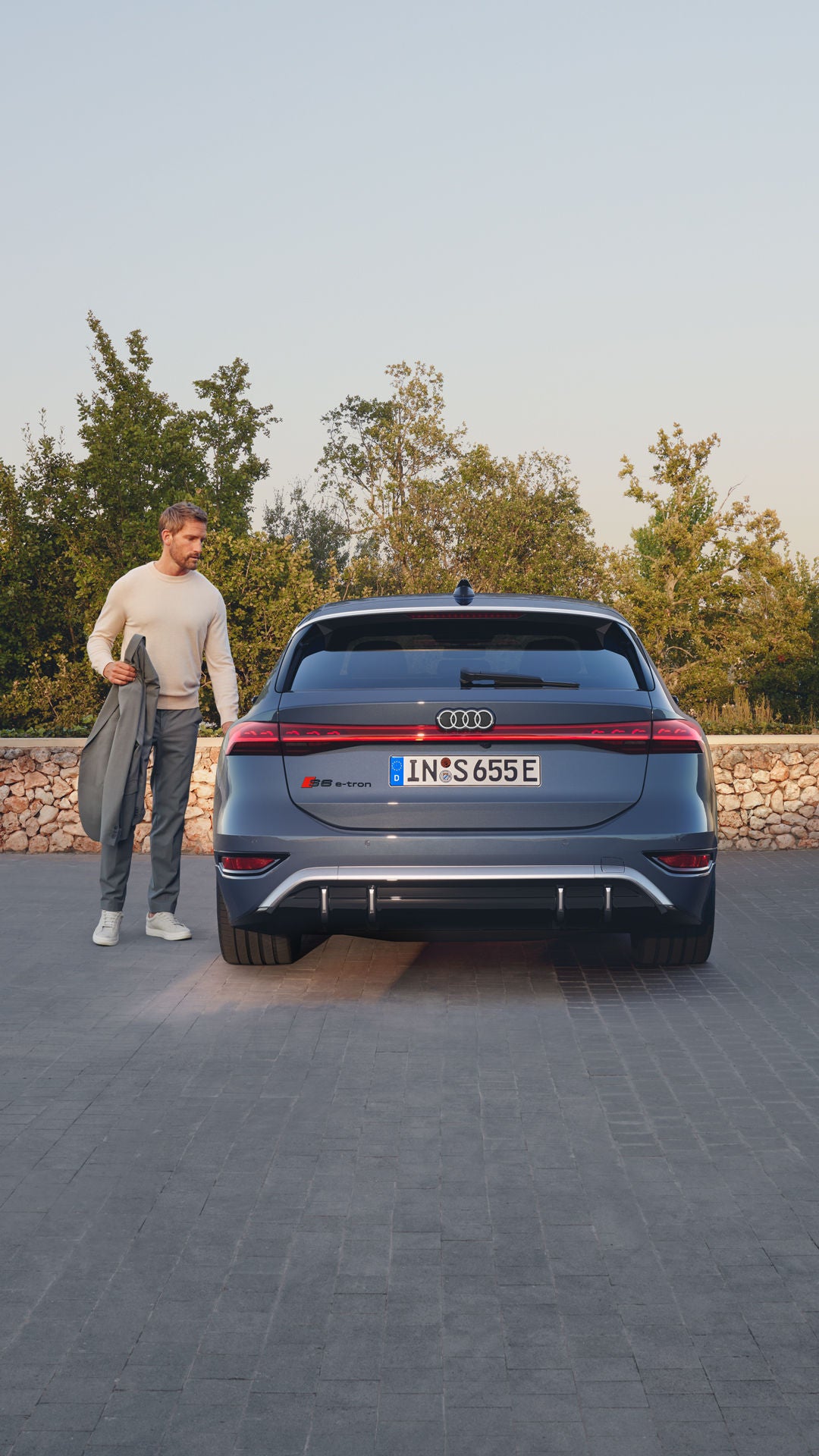 Rückansicht eines grauen Audi. An der Fahrertür steht ein blonder Mann mit einer grauen Jacke in der Hand. Er scheint kurz davor, in das Auto einzusteigen.