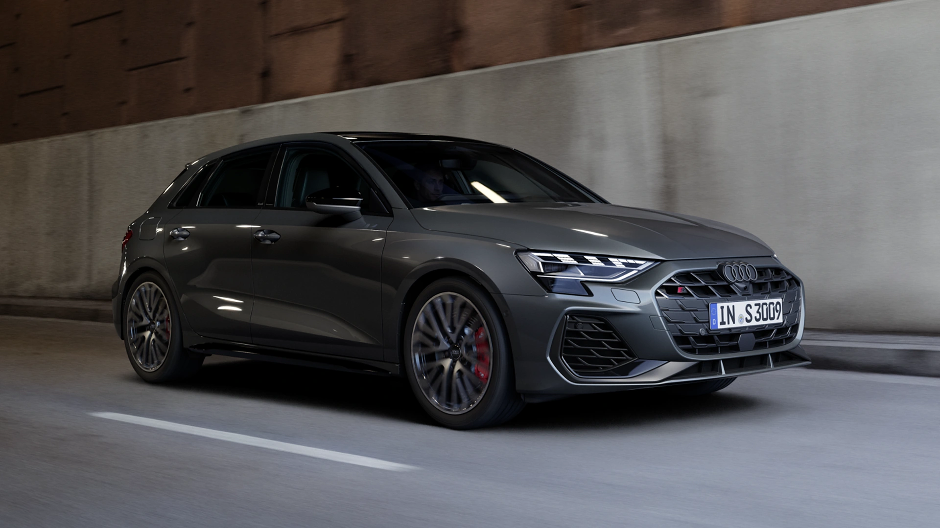 A grey hatchback car parked at an urban location with a concrete building backdrop.