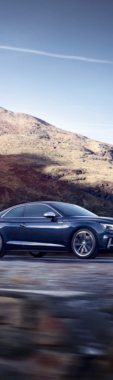 Dunkelblauer Audi S5 fährt auf Bergstraße, im Hintergrund felsige Landschaft mit sonnigem Gipfel unter klarem Himmel.