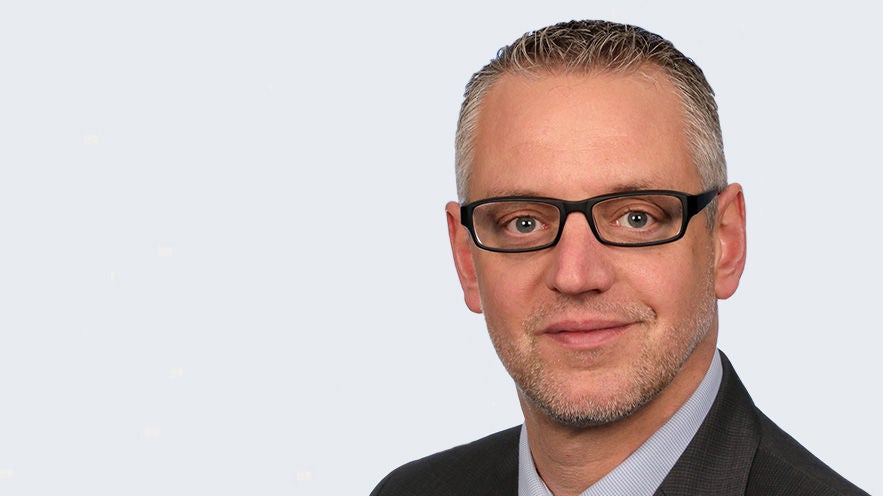 Thomas Eberleh: Porträt Mann mit grauem Kurzhaarschnitt und schwarzer Brille im Anzug vor grauem Hintergrund.