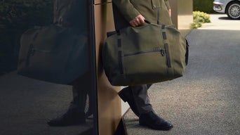 Person trägt große olivgrüne Audi-Reisetasche vor Gebäudeeingang, im Hintergrund steht ein silbernes Auto.