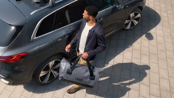 Mann mit grauer Audi Outdoor-Reisetasche steht neben dunklem SUV auf gepflastertem Platz.
