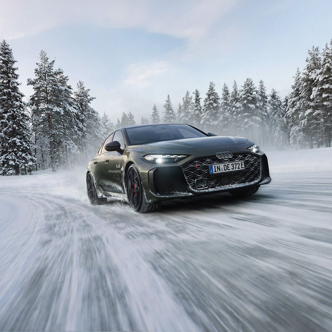 A dark green Audi RS 5 sedan drives along a snow-covered road; snow dust, a pine forest, and a blue sky.