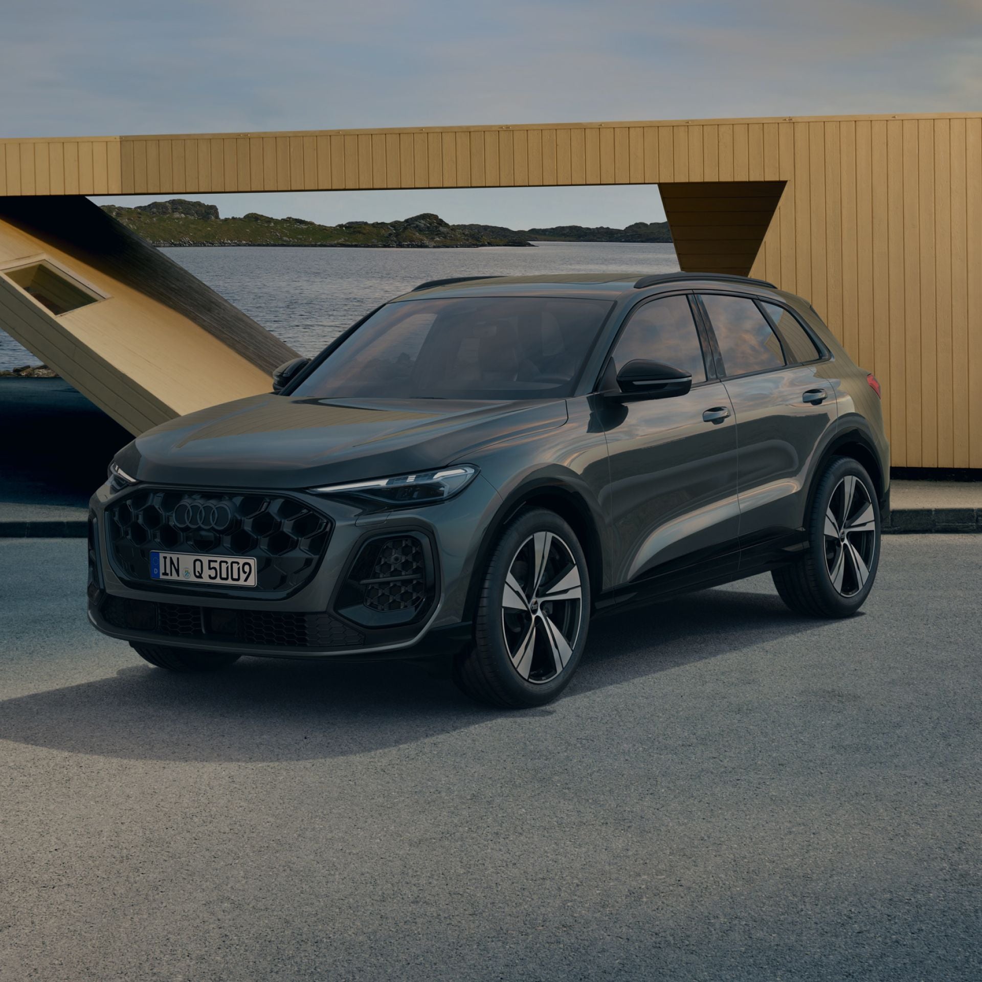 A grey Audi Q5 Sportback TFSI e parked by a scenic waterfront, with modern architectural elements in the background under a clear sky.