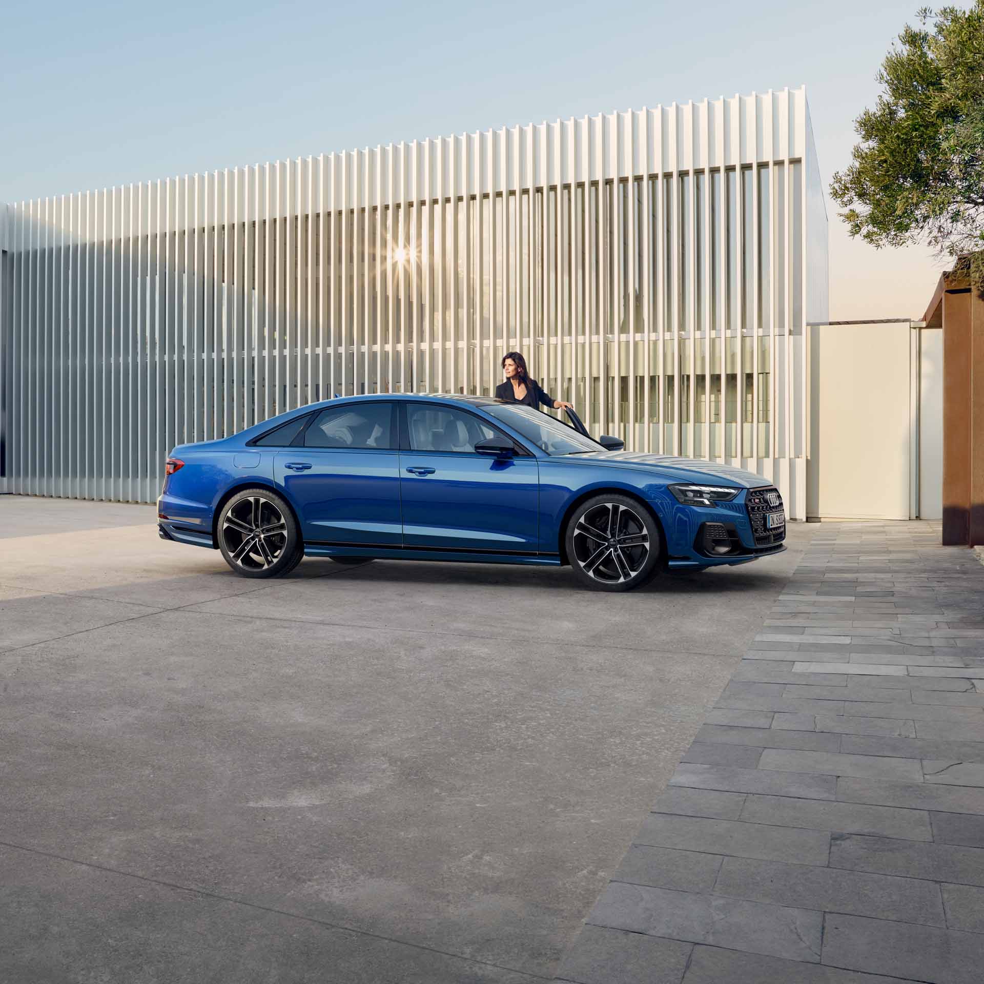 Ein blauer Audi S8 TFSI steht vor einer Wand mit vertikalen Metalllamellen, dunkelhaarige Frau steigt ein.